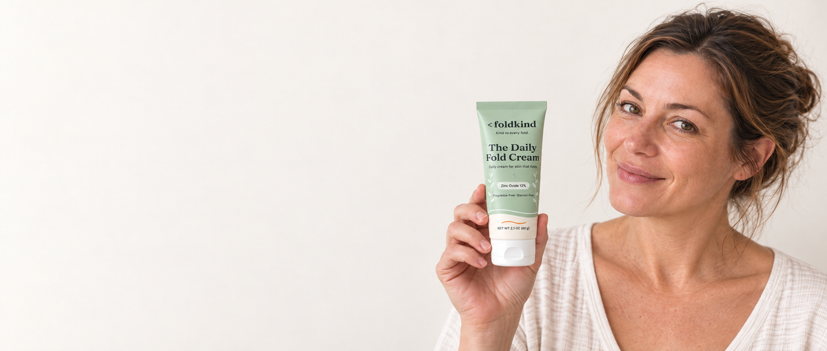 A woman holding The Daily Fold Cream against a plain ivory wall in morning light. Daily cream for skin that folds, zinc oxide 12%, 2.1 oz (60 g).