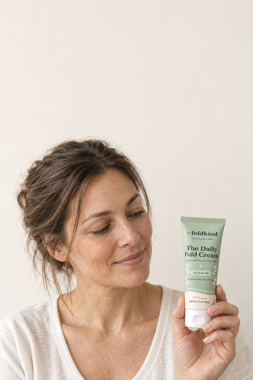 A woman holding The Daily Fold Cream against an ivory wall in morning light, the label showing zinc oxide 12%, fragrance-free and steroid-free, 2.1 oz (60 g).